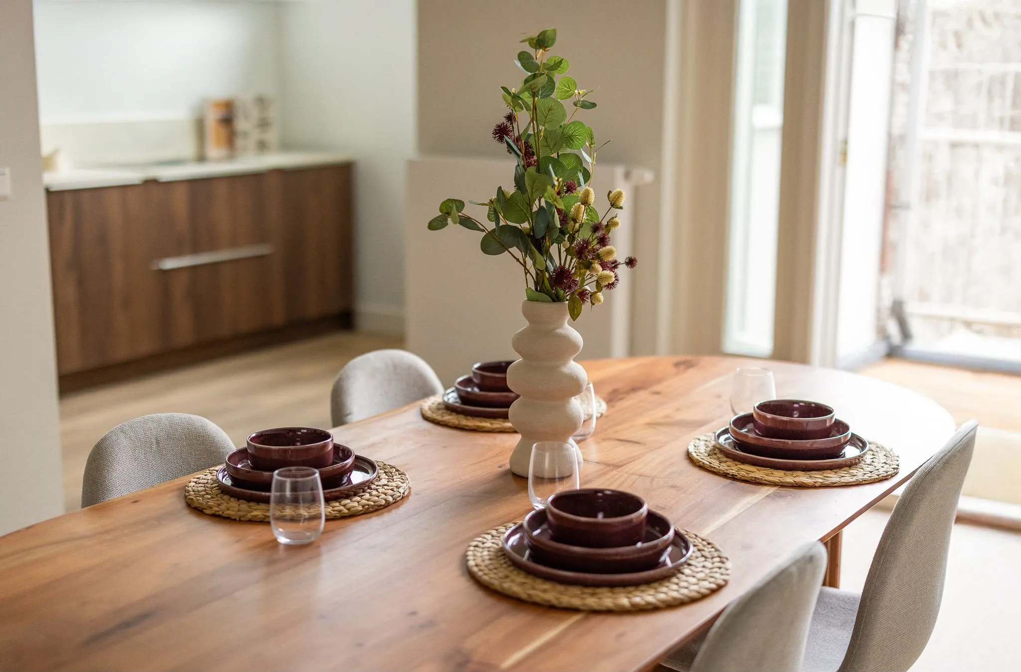 Houten eettafel gedekt met aubergine servies op rieten onderzetters, een ribbelvaas met bloemen en de notenhouten keuken op de achtergrond