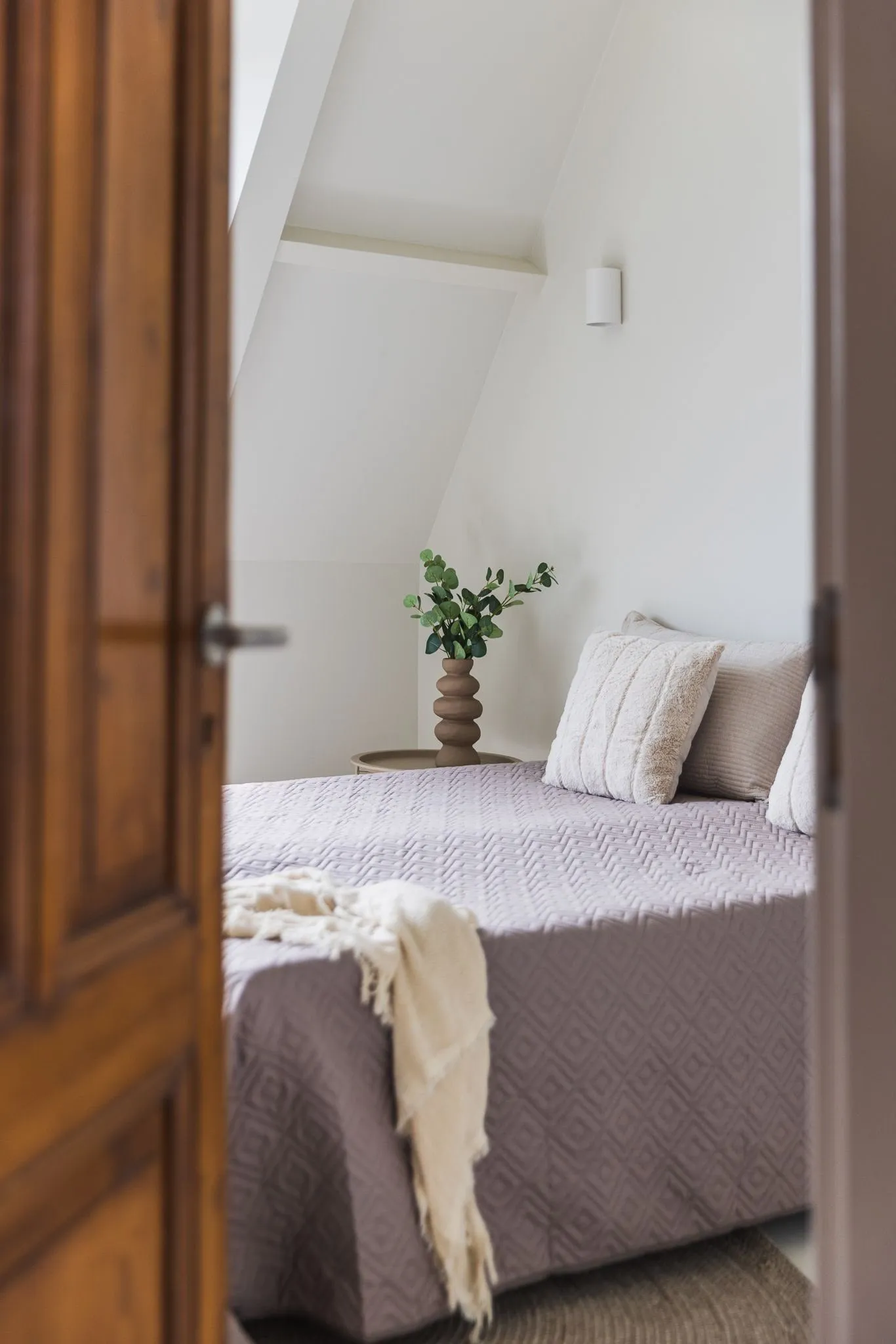Doorkijk door een houten deur naar een slaapkamer onder een schuin dak met een lila sprei en een gebreide plaid