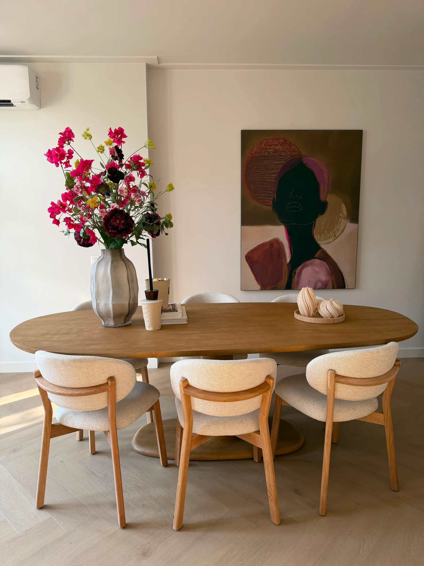 Ovale houten eettafel met bouclé stoelen, een groot boeket met roze bloemen en een portretschilderij aan de muur